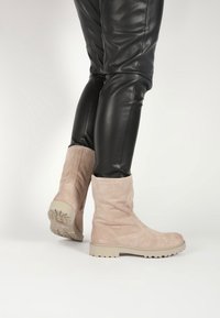 Botines de ante de color beige claro con un diseño sencillo, suelas de goma contrastantes y una textura suave; combinados con pantalones de cuero negros.