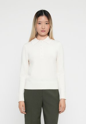 Polo blanc à manches longues avec un ourlet côtelé, doté d'un col et d'une patte de boutonnage, porté avec un pantalon noir vert ajusté.