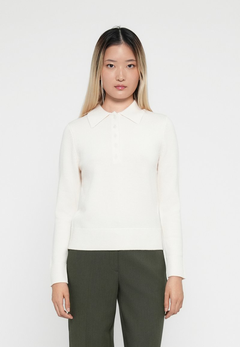 Bílý svetr s dlouhým rukávem a polo límcem, ribbovaným lemem, s knoflíkovou légou, nošený s tmavě zelenými elegantními kalhotami.