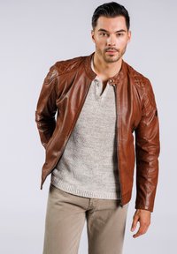 Bruine leren jas met gequiltte schouderaccenten, gecombineerd met een lichtgrijze gebreide trui en beige broek. Ritsluiting aan de voorkant.