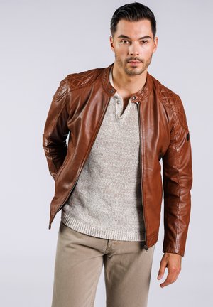 Chaqueta de cuero marrón con acentos acolchados en los hombros, combinada con un suéter de punto gris claro y pantalones beige. Cierre con cremallera en la parte frontal.