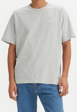 Homme portant un t-shirt Levi's gris clair uni et un jean bleu, debout devant un fond blanc.
