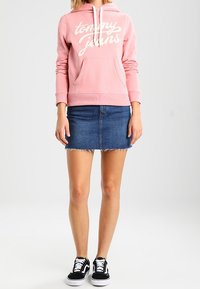 Sudadera rosa claro con logo de "Tommy Jeans", bolsillo y cordones, combinada con una falda mini de mezclilla y zapatillas negras.