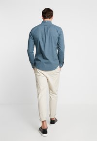 Blå skjorta med knappar, mjukt tyg, långa ärmar och rundad nederkant, i kombination med ljusbeiga byxor och svarta slip-on skor.