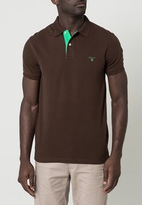 Polo marron avec des accents verts sur le col, patte à deux boutons, et logo brodé sur la poitrine. Manches courtes et tissu en coton.