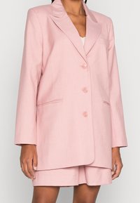 Světle růžový blazer vyrobený z texturované látky, s klopovým límcem, třemi předními knoflíky a dvěma bočními kapsami pro praktické použití.