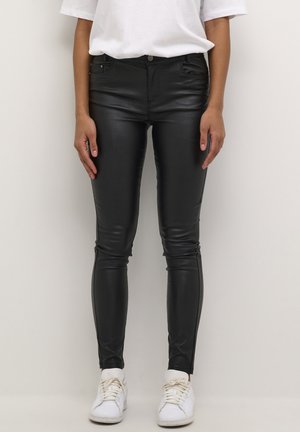 Schwarze, schmal geschnittene Jeans aus Kunstleder mit hoher Taille, glatter Textur und frontalen Taschen, kombiniert mit weißen Sneakers und einem lockeren weißen T-Shirt.