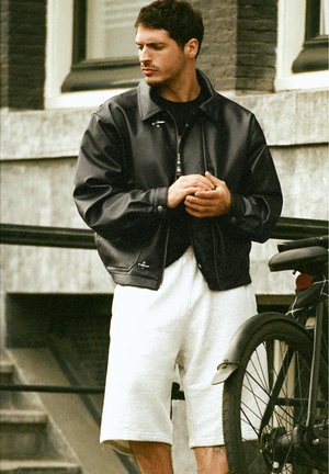 Mann in schwarzer Lederjacke und weißen Shorts steht im Freien neben einem schwarzen Fahrrad in urbaner Umgebung und schaut zur Seite.