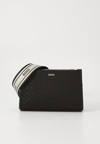 Borsa rettangolare nera in materiale testurizzato con logo dorato. Presenta una tracolla bianca a contrasto con scritte nere. Chiusura con zip.