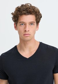 Młody mężczyzna o kręconych brązowych włosach i niebieskich oczach, ubrany w czarny t-shirt z dekoltem w serek, na jednolitym jasnym tle.