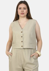 Beige mouwloos vest met een diepe V-hals, vijf bruine knopen en zijzakken, gemaakt van gestructureerde stof. Gestyleerd met losse broek.