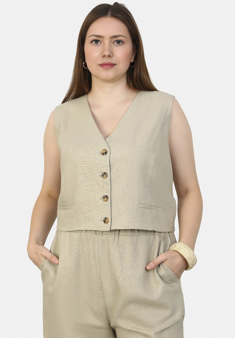 Beige mouwloos vest met een diepe V-hals, vijf bruine knopen en zijzakken, gemaakt van gestructureerde stof. Gestyleerd met losse broek.