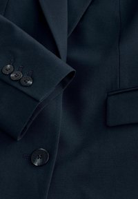 Blazer bleu marine foncé avec revers cranté, quatre boutons noirs aux poignets, un bouton noir sur le devant et une poche à rabat sur le côté droit.