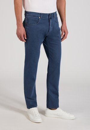 Man draagt marineblauwe rechte broek met voor- en achterzakken, gecombineerd met witte lage vetersneakers.