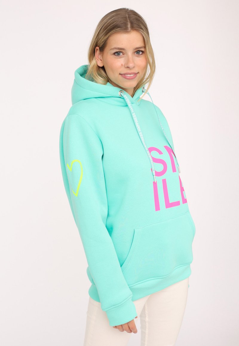 Mint green hoodie with pink "SMILE" text, yellow heart detail on left sleeve, large front pocket, and drawstring hood. Soft fabric texture.