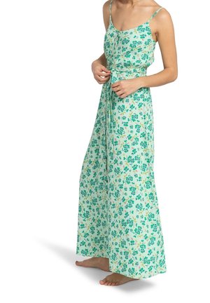 Femme ajustant la ceinture d’une robe sans manches, longue jusqu’au sol, avec un motif floral vert et bleu, debout pieds nus sur fond blanc.