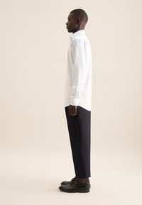 Camicia a righe verticali bianche con maniche lunghe, abbinata a pantaloni tapered blu scuro e scarpe in pelle nere, vista di lato.