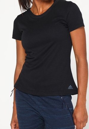 Vrouw draagt een aangesloten zwart Adidas T-shirt en marineblauwe sportbroek met rits en staat tegen een effen witte achtergrond.