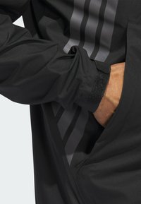 Svart jacka med grå ränder, tillverkad av ett slätt, texturerat material. Inkluderar justerbara kardborreband vid ärmsluten och en sidoficka.