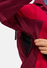 Bordeaux jakke med en lynlåslomme på siden. Stoffet ser glat og let blankt ud, med kontrastfarvede røde paneler.