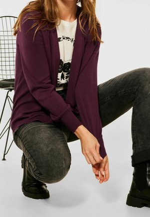 Cardigan violet à front ouvert, manches longues et poignets côtelés, porté par-dessus un t-shirt graphique noir et blanc. Jean foncé et bottines noires.