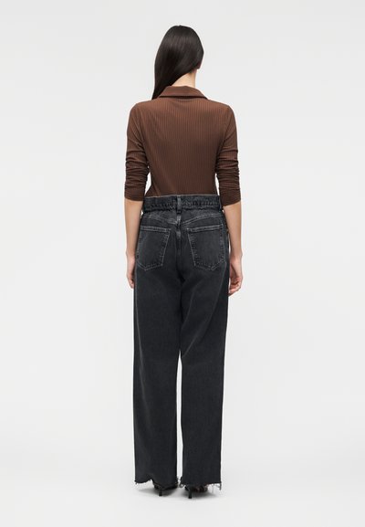 Γυναίκα με μακριά σκούρα μαλλιά, που φοράει ένα καφέ ριμπ μακρυμάνικο τοπ και μαύρο, φαρδύ τζιν με ψηλή μέση και ξεφτισμένα τελειώματα, που στέκεται με την πλάτη γυρισμένη.