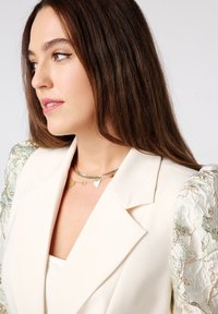 Stijlvolle witte blazer met gestructureerde, gemusterde mouwen, gelaagde gouden hartketting en glad haar dat het gezicht omlijst. Schone achtergrond.