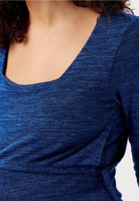 Kobieta ubrana w dopasowaną, długosleeve, niebieską bluzkę o heatherowanym wzorze z kwadratowym dekoltem i delikatnym marszczeniem w okolicy talii.