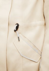 Veste beige présentant un tissu texturé, une poche latérale avec des surpiqûres contrastantes, un cordon de serrage avec embout, et une doublure intérieure lisse.