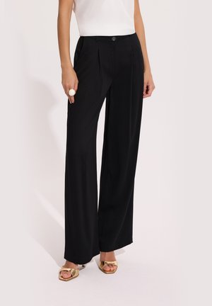 Kobieta ubrana w czarne szerokie spodnie, białą bezrękawnik, złote sandały na obcasie z odkrytymi palcami oraz dużą białą obrączkę na lewej dłoni.