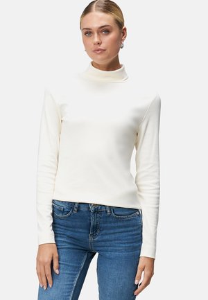 Haut à col roulé crème à manches longues en matière douce, design ajusté, texture lisse, porté avec un jean bleu foncé.