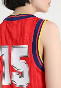 Maillot de basketball rouge avec le numéro blanc "15", présentant une texture en maille, un col rayé bleu et jaune, et un design sans manches.