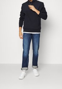 Sudadera azul marino con cuello alto, puños acanalados y bolsillos laterales, combinada con vaqueros de mezclilla azul claro y zapatillas blancas con detalles en rojo.