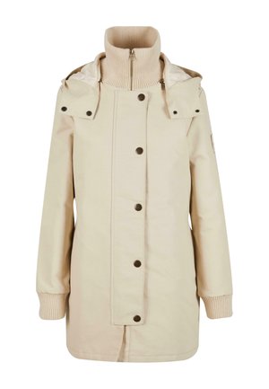 Beige jakk, millel on kõrge soonikkoes kaelus, kinnitusnupud ja eemaldatav kapuuts. Omab küljekotte ja soonikkoes varrukaotsi. Sile kangateks.