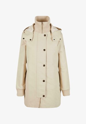 Beige jakk, millel on kõrge soonikkoes kaelus, kinnitusnupud ja eemaldatav kapuuts. Omab küljekotte ja soonikkoes varrukaotsi. Sile kangateks.