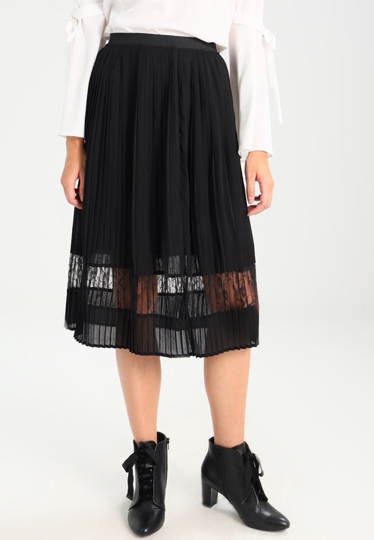 Femme portant une jupe midi plissée noire avec un détail en dentelle transparente à l'ourlet, un haut blanc à manches longues et des bottines noires à lacets avec talons.