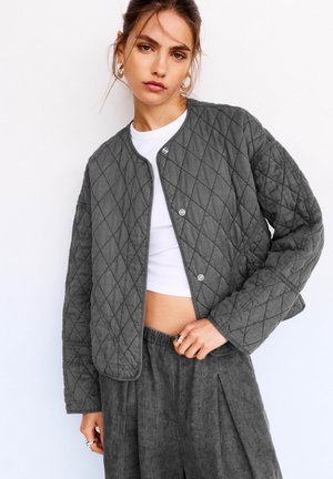 Grau gesteppte Jacke mit Druckknöpfen, runden Ausschnitt und herunterfallenden Schultern, kombiniert mit einem weißen Oberteil und strukturierten grauen Hosen.