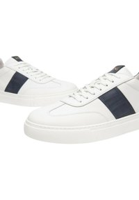 Sneakers bianchi in pelle con una striscia diagonale blu scuro, punta tonda e suole in gomma testurizzata, lacci piatti e branding dorato.