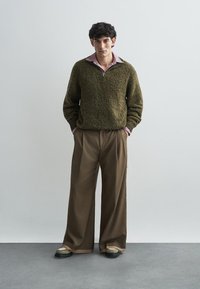 Pulover texturat verde măsliniu cu guler cu fermoar, asortat cu pantaloni maro, evazați, și pantofi negri masivi. Cămașă roz deschis dedesubt.