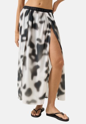 Kobieta nosząca czarno-białą wzorzystą maksikę z wysokim bocznym rozcięciem, czarnym elastycznym pasem i czarnymi klapkami.