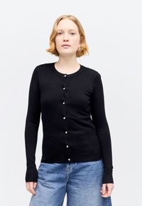 Νεαρός ενήλικας με κοντά ξανθά μαλλιά, φοράει μαύρη ζακέτα με κουμπιά και μπλε τζιν παντελόνια, στέκεται μπροστά σε απλό φωτεινό φόντο.