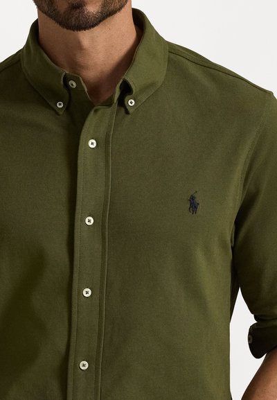 Camisa de colarinho verde escuro feita de tecido texturizado, com botões brancos e um pequeno logótipo azul-marinho no lado esquerdo.