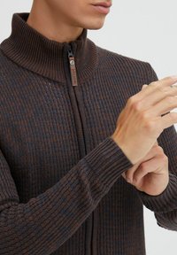 Mørk ribbet sweater med høj krave, præsenterer en lynlås foran og en struktureret strikket mønster i brune og grå nuancer.
