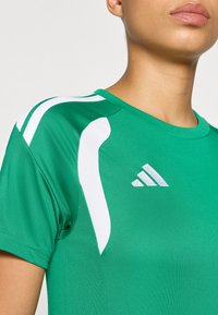 Πράσινο αθλητικό πουκάμισο με λευκές λεπτομέρειες, με σ τρογγυλή λαιμόκοψη και δίχτυ στη μπροστινή πλευρά του ώμου. Το λογότυπο της Adidas εμφανίζεται στο μπροστινό μέρος.