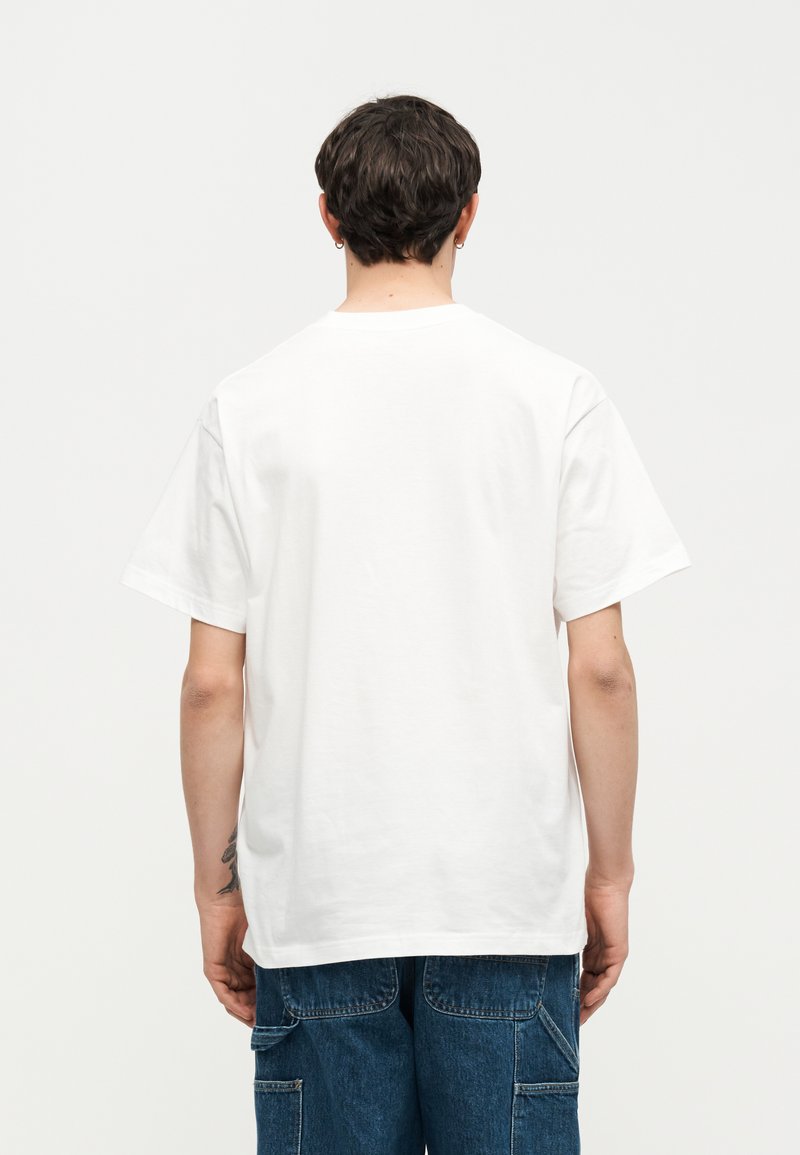 Uomo con capelli corti e scuri che indossa una maglietta bianca semplice a maniche corte e jeans blu, visto da dietro su sfondo bianco.