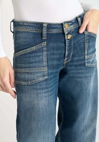 Jeans de mezclilla azul con un lavado deslavado, que cuentan con dos bolsillos delanteros, un cierre de doble botón y detalles de costura intrincados.