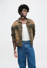 Καφέ τζιν μπουφάν με φθαρμένη υφή, εμπρόσθιες τσέπες και ασημένια κουμπιά, φορεμένο πάνω από λευκό τοπάκι και μπλε τζιν ψηλής μέσης.