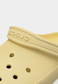 Κίτρινο παπούτσι Crocs με οπές αερισμού και ανάγλυφο λογότυπο στο ρυθμιζόμενο λουράκι της φτέρνας σε λευκό φόντο.