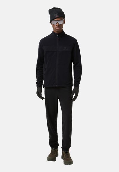 Black fleece zip-up jacket with a horizontal panel, black gloves, and pants. Worn with a black beanie and reflective sunglasses.