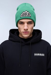 Berretto a maglia verde con un logo di montagna e testo ricamato in nero; abbinato a una felpa con cappuccio nera con un piccolo logo sul petto.